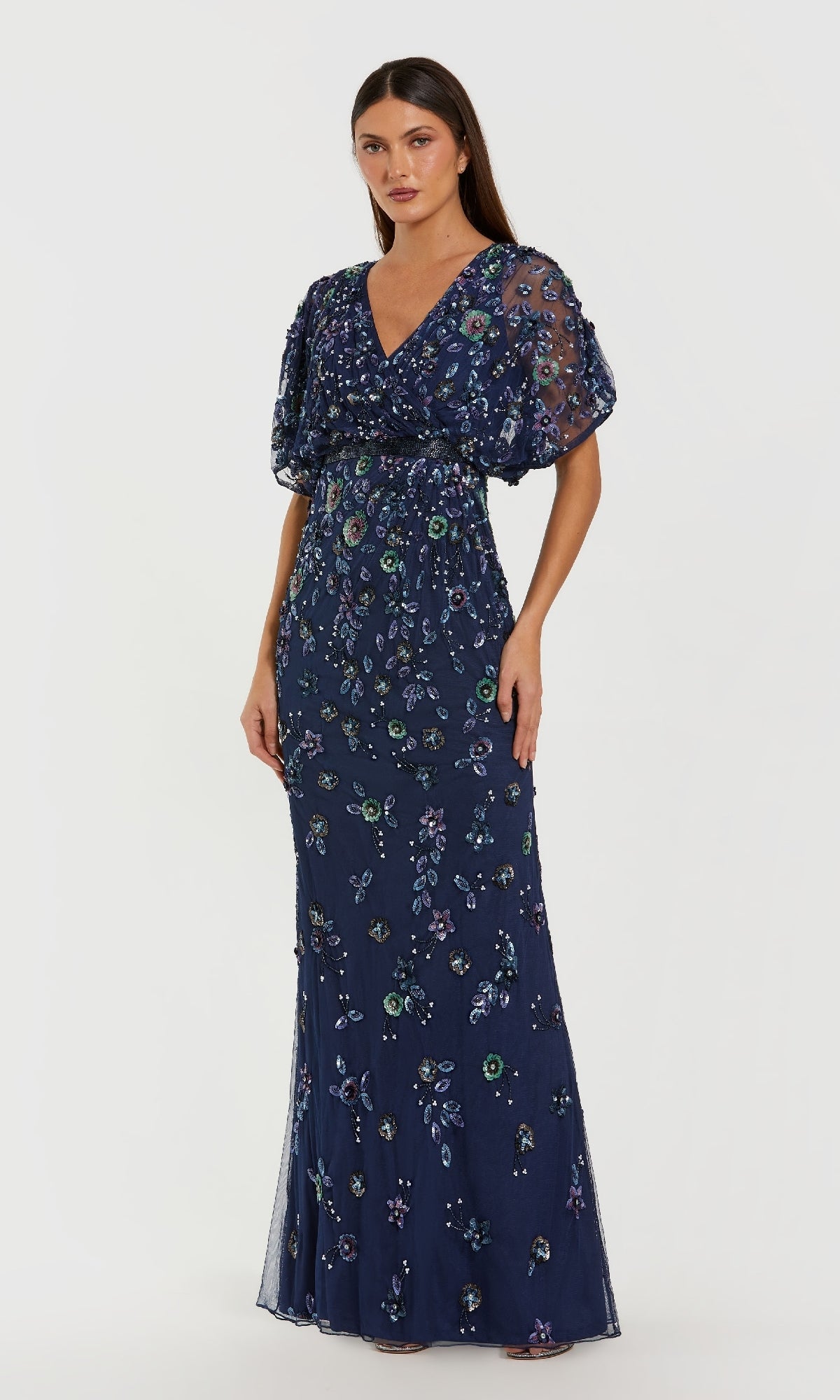 A woman wears the Mac Duggal 6158 Short Sleeve Beaded Formal Dress, a floor-length gown with sheer sleeves and multicolored floral embroidery, set against a plain white background.